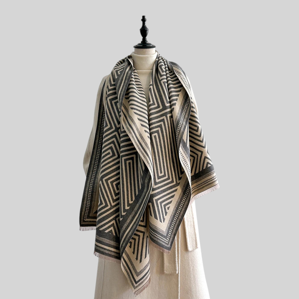 A large woven shawl with a geometric maze-like pattern in beige and dark gray, draped over a cream-colored dress form against a solid light gray background.