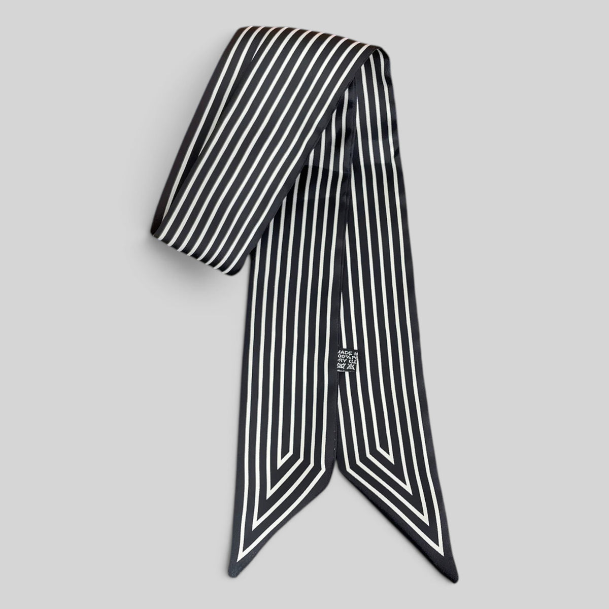 A black silk scarf with white vertical stripes and a small silver-toned logo badge, displayed on a neutral gray background.