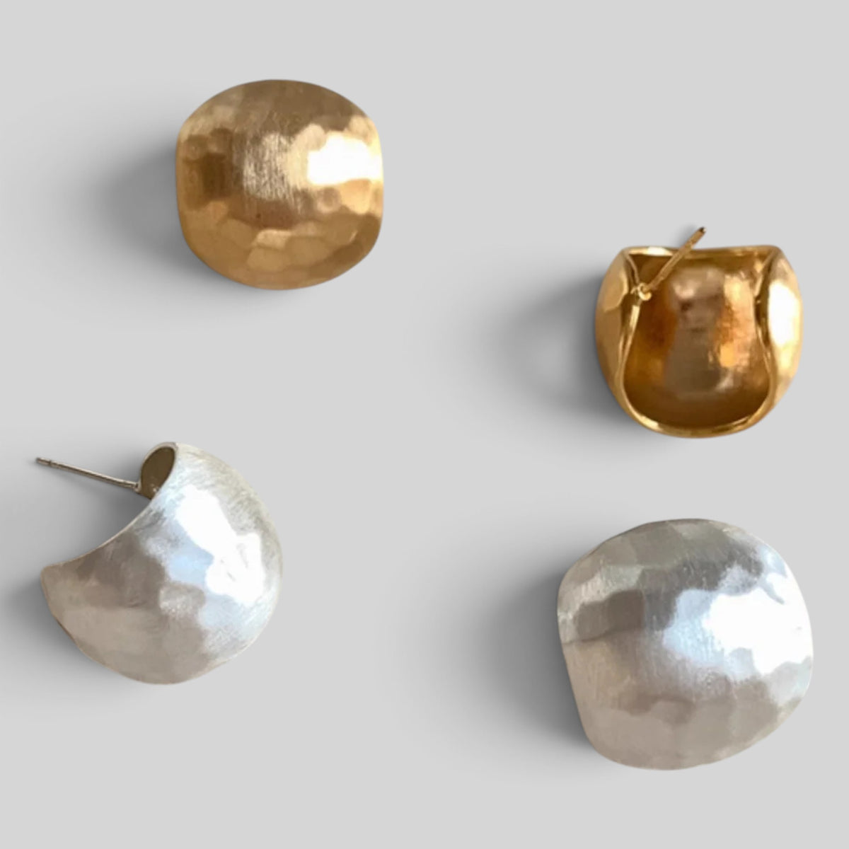 Four sculptural earrings with organic, hammered finishes in gold and silver, arranged on a neutral light gray background.