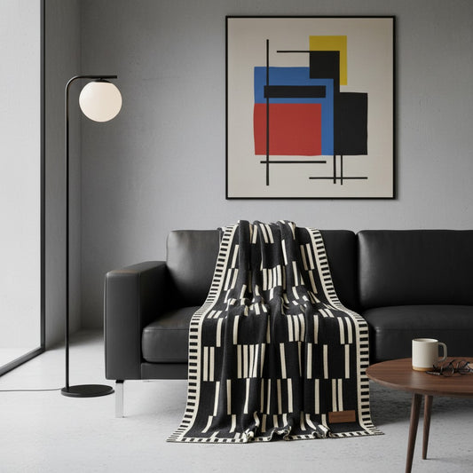 A modern living room scene featuring a black leather sofa draped with a black and cream geometric patterned throw blanket. A black floor lamp stands to the left, and a round wooden coffee table with a white mug is in the foreground.