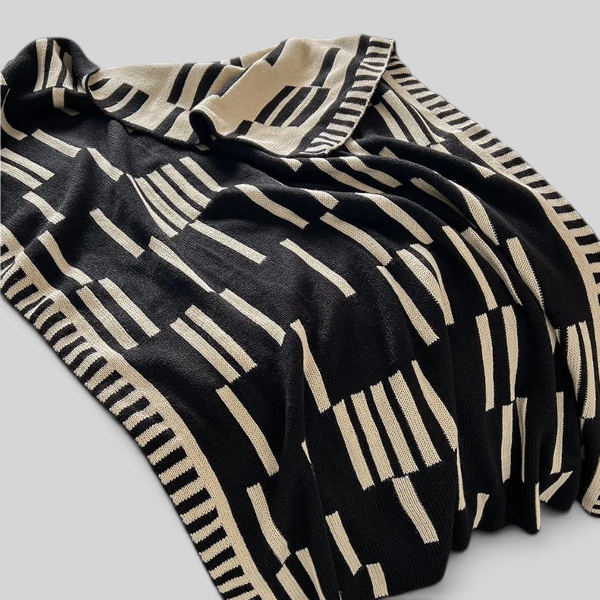 A black knit blanket with a cream-colored geometric stripe pattern, draped on a light gray background.