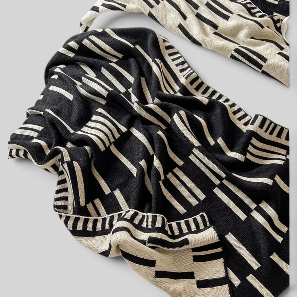 A draped knit scarf featuring a bold black and cream geometric stripe pattern, shown against a neutral light gray background.