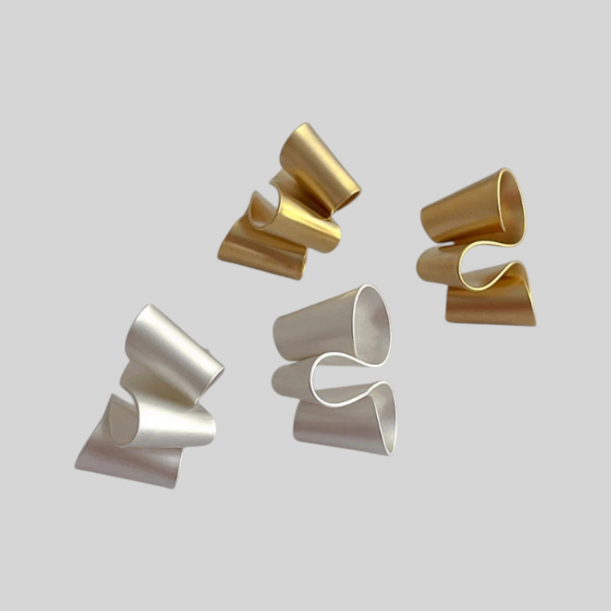 Four sculptural, curved metallic rings in polished gold and silver finishes, arranged on a solid light gray background.