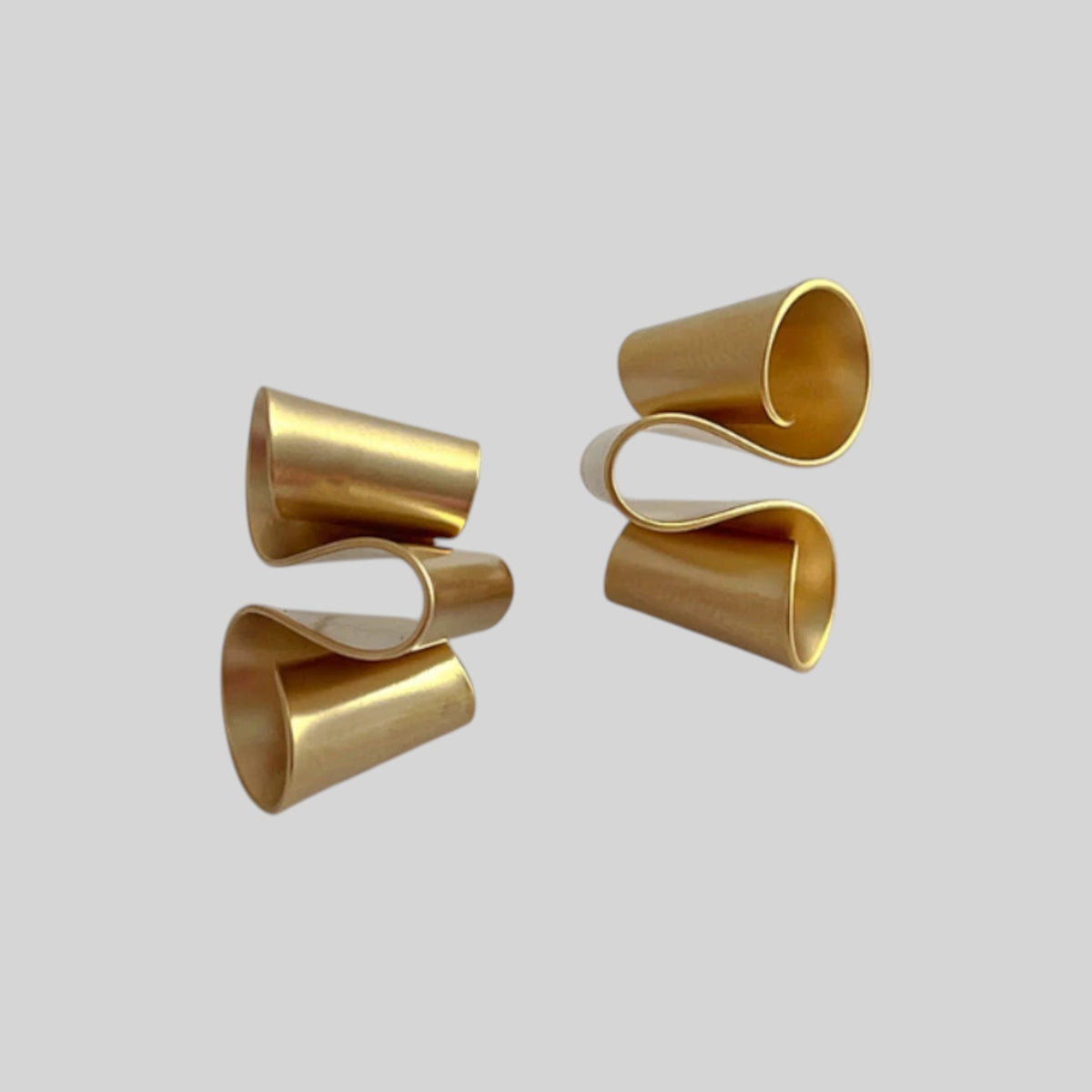 A pair of sculptural gold-toned earrings with a wavy, curled tube design, displayed on a solid light gray background.