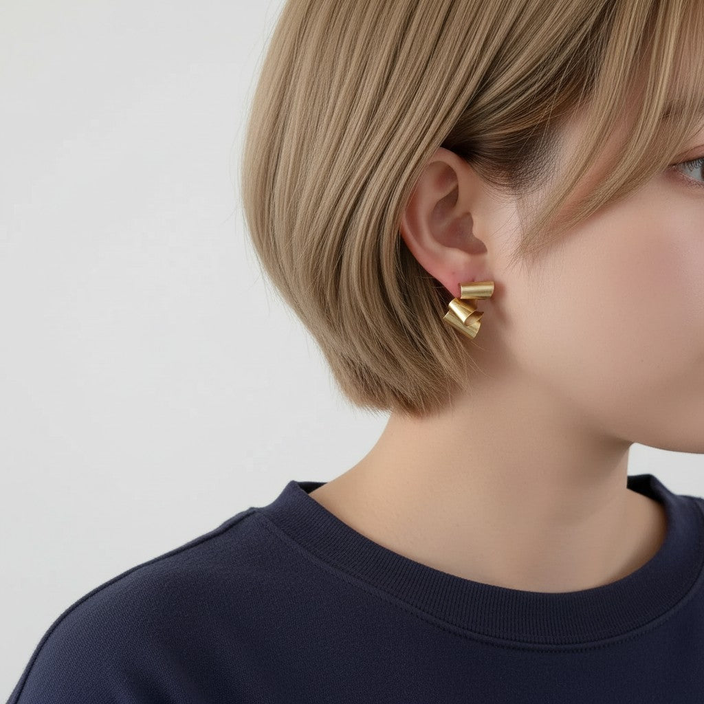 A young woman with a light brown bob, wearing a blue sweater and matte gold geometric earrings on a solid light gray background
