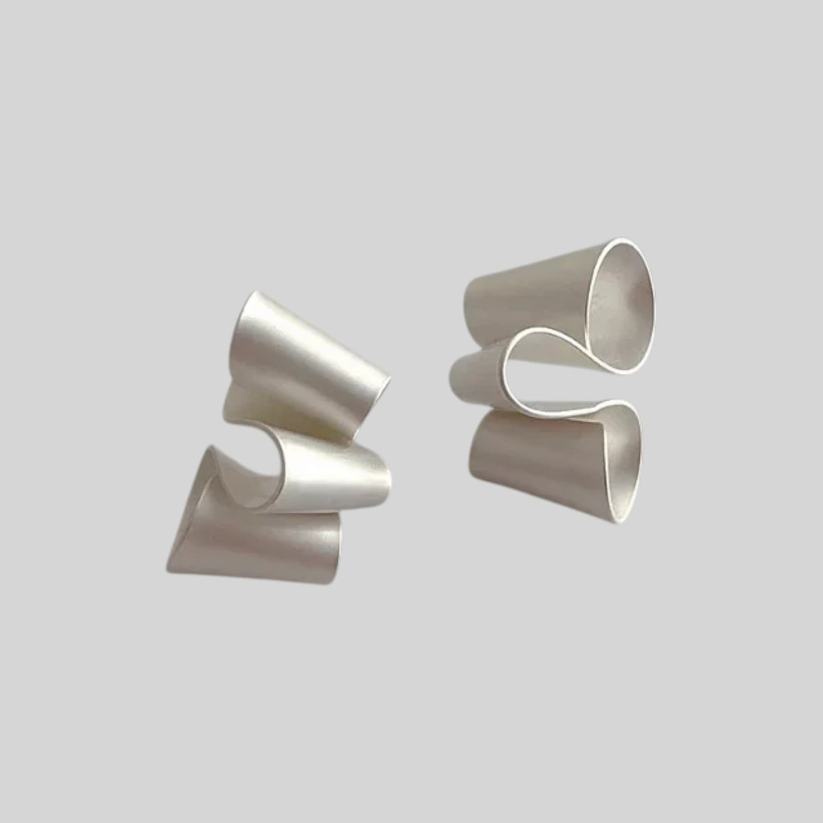 A pair of sculptural, silver-toned earrings with a wavy, layered design, displayed against a solid light gray background.