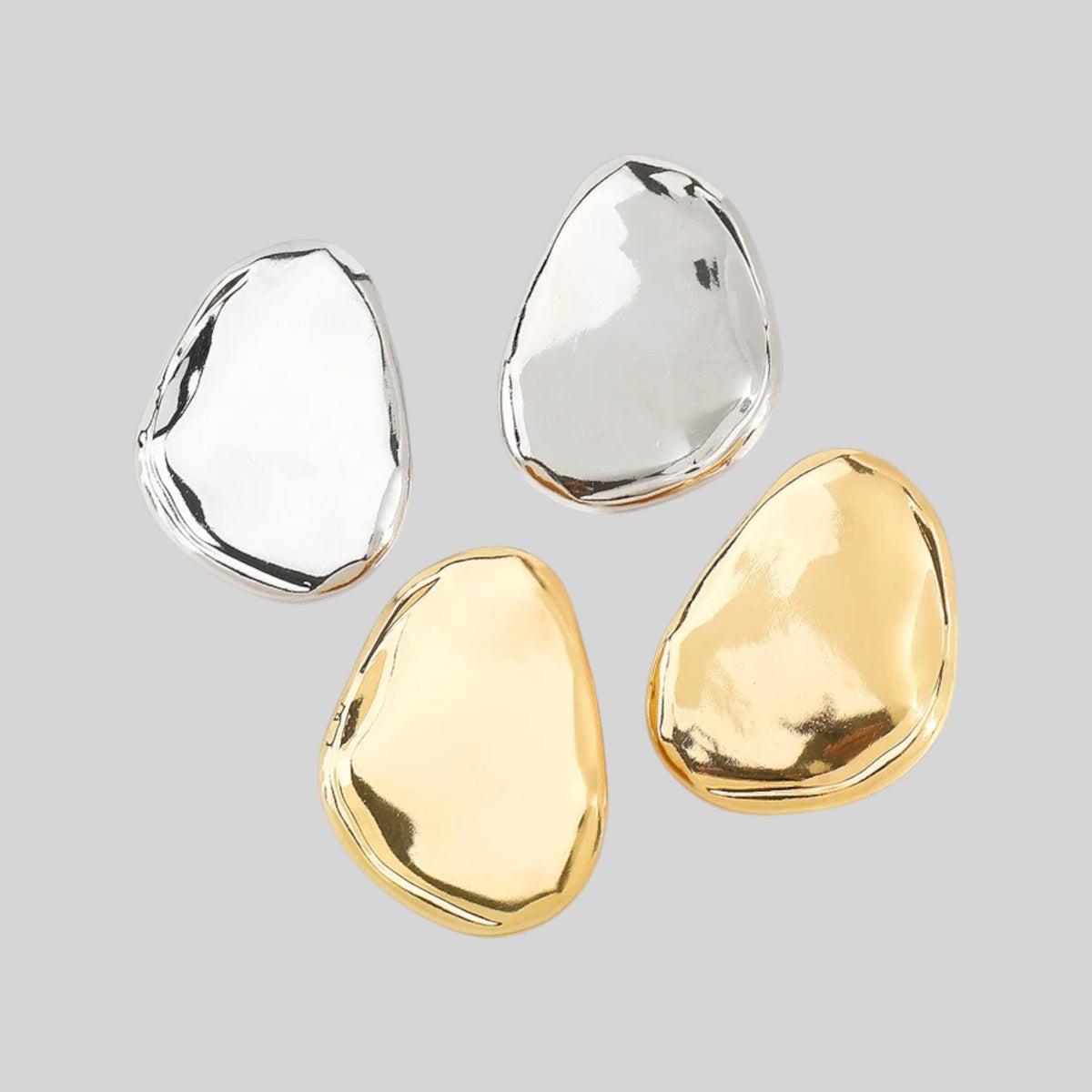Four irregular, polished stone-like jewelry pieces in silver and gold finishes, arranged on a solid light gray background.