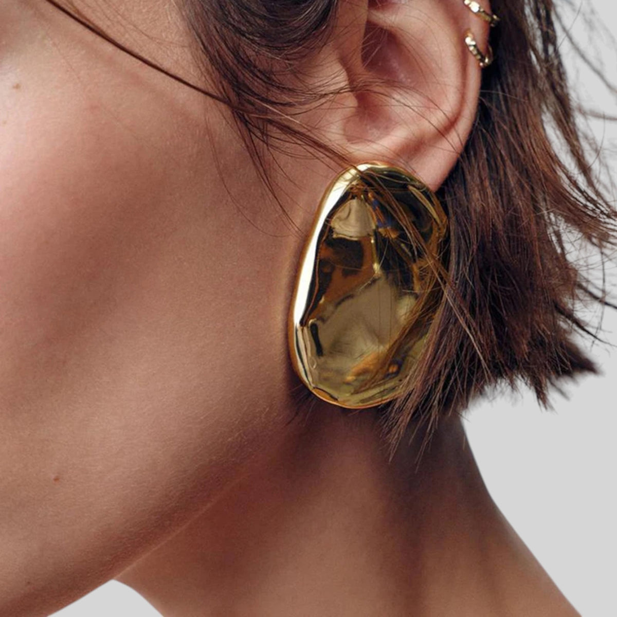 A close-up of a person's ear wearing a large, organic-shaped gold earring with a highly reflective, polished metallic surface.