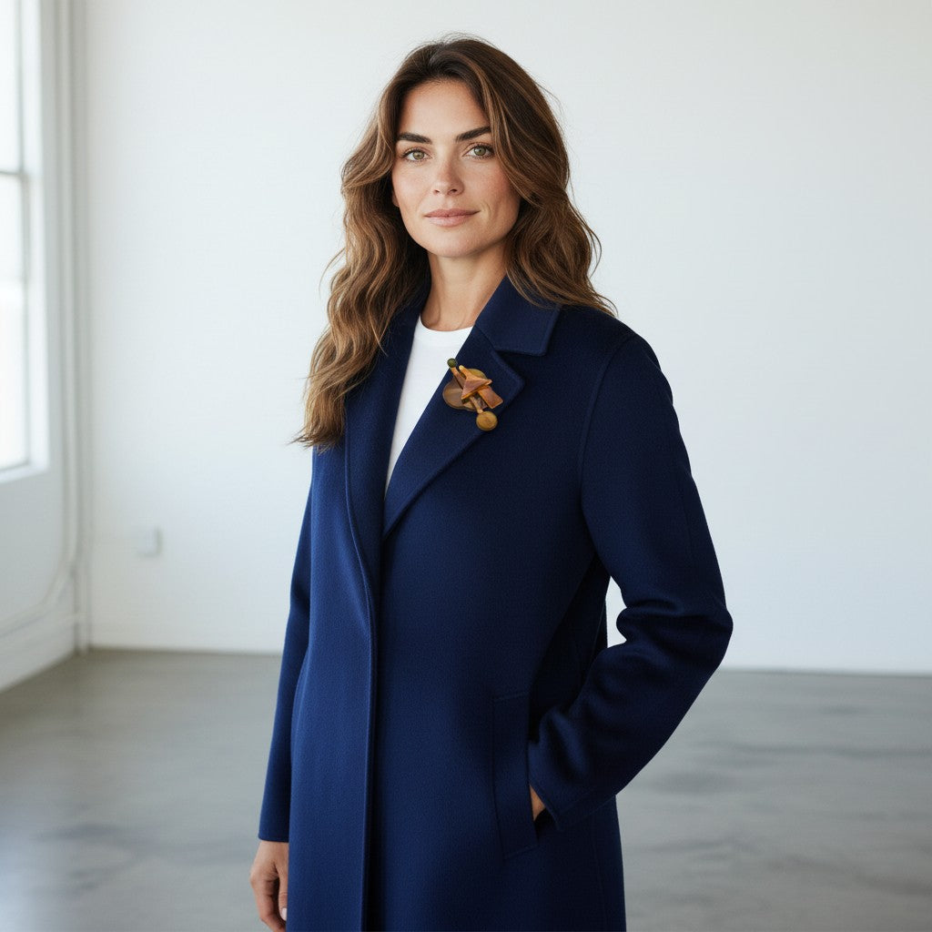 A woman wearing a navy blue wool-blend coat over a white top, accessorized with a brown floral brooch, standing in a bright, minimalist room with white walls and gray concrete floors.