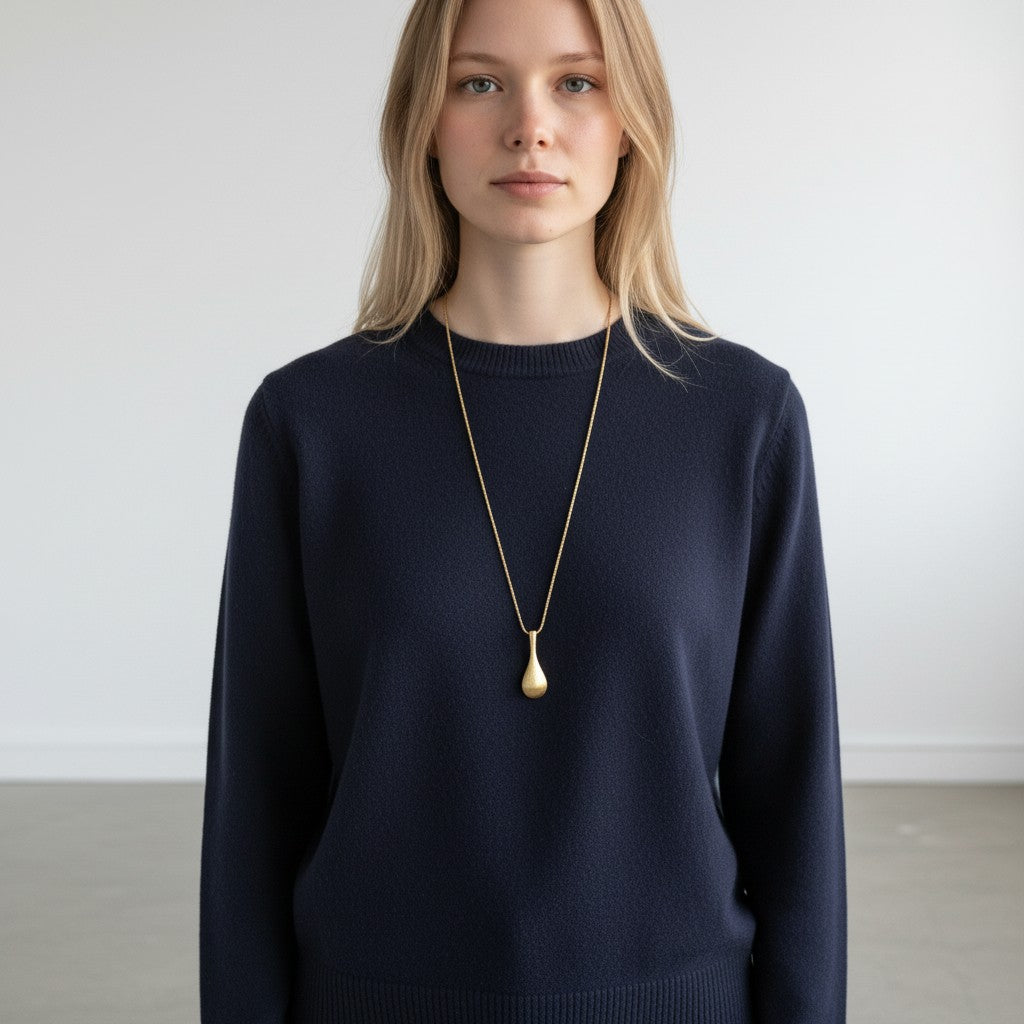 A blonde woman wearing a dark navy blue knit sweater and a long gold pendant necklace with a teardrop-shaped drop, standing against a plain light gray wall.