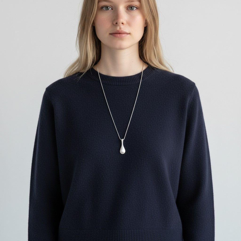 A woman wearing a navy blue knit crewneck sweater and a long silver pendant necklace with a teardrop-shaped charm, photographed against a light gray studio background.