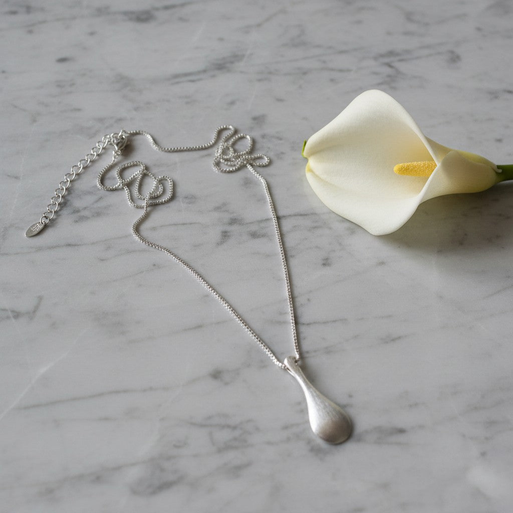 A silver teardrop pendant necklace with a script-style chain spelling 'love', displayed on a white and gray marble surface next to a white calla lily flower.