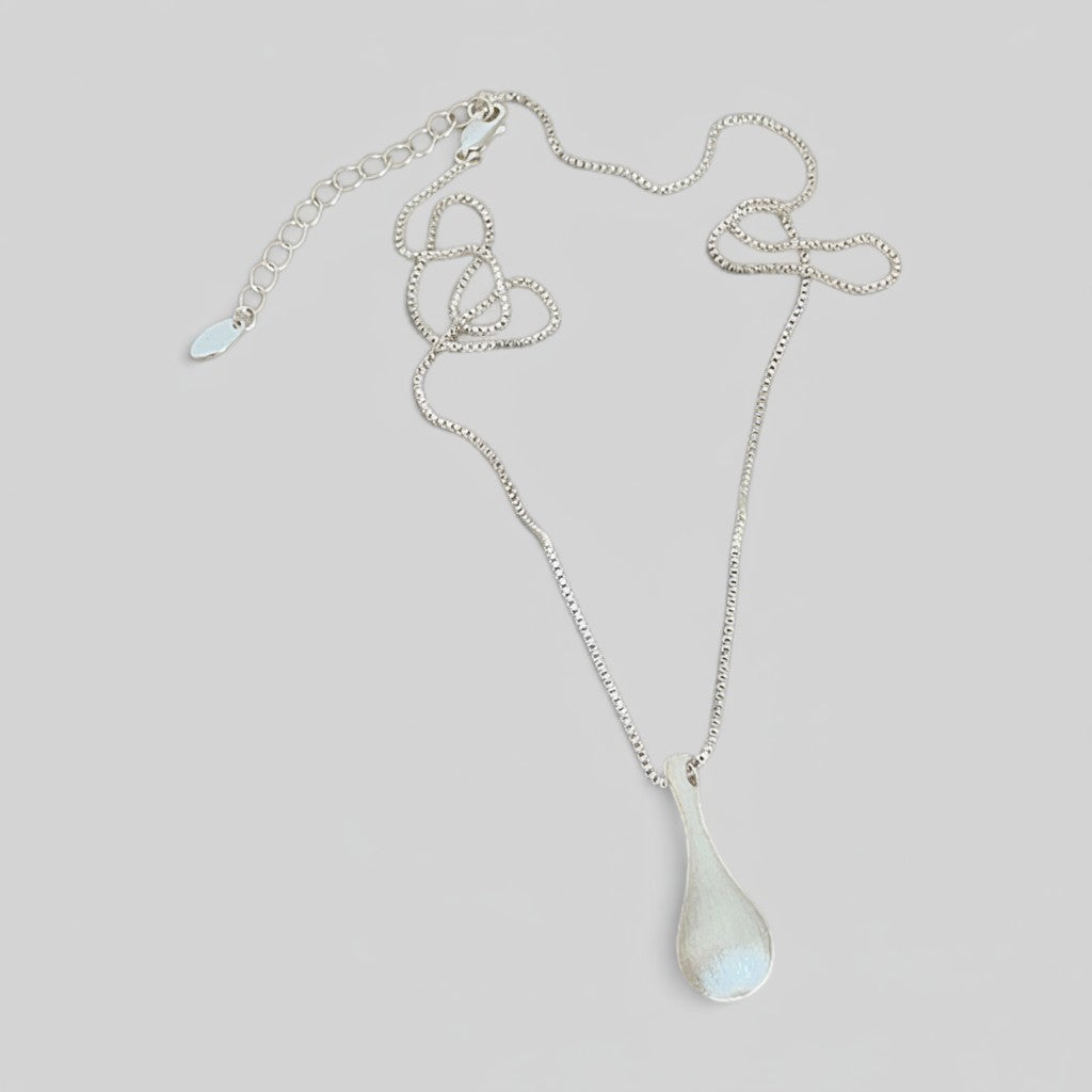 A silver chain necklace with a teardrop-shaped pendant featuring a white iridescent inlay, displayed on a neutral light gray background.