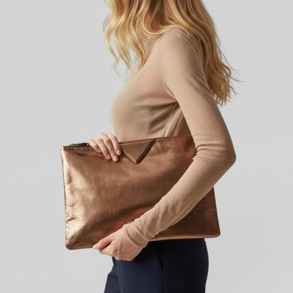 A woman in a beige long-sleeve top and navy pants holds a metallic rose gold envelope-style clutch bag against a light gray studio background.