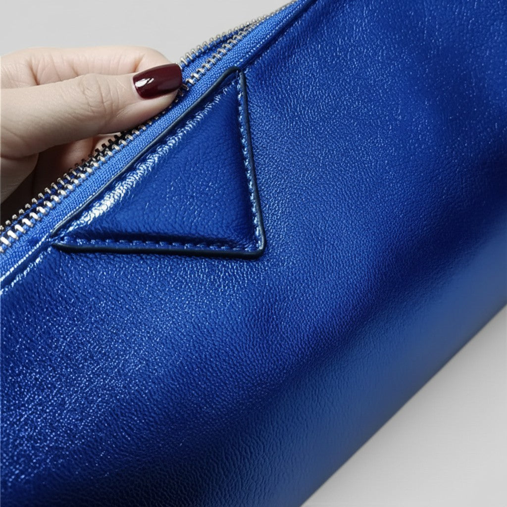A close-up of a metallic royal blue leather pouch with a hand sliding a silver zipper tab, showing a triangular stitched detail on the front.