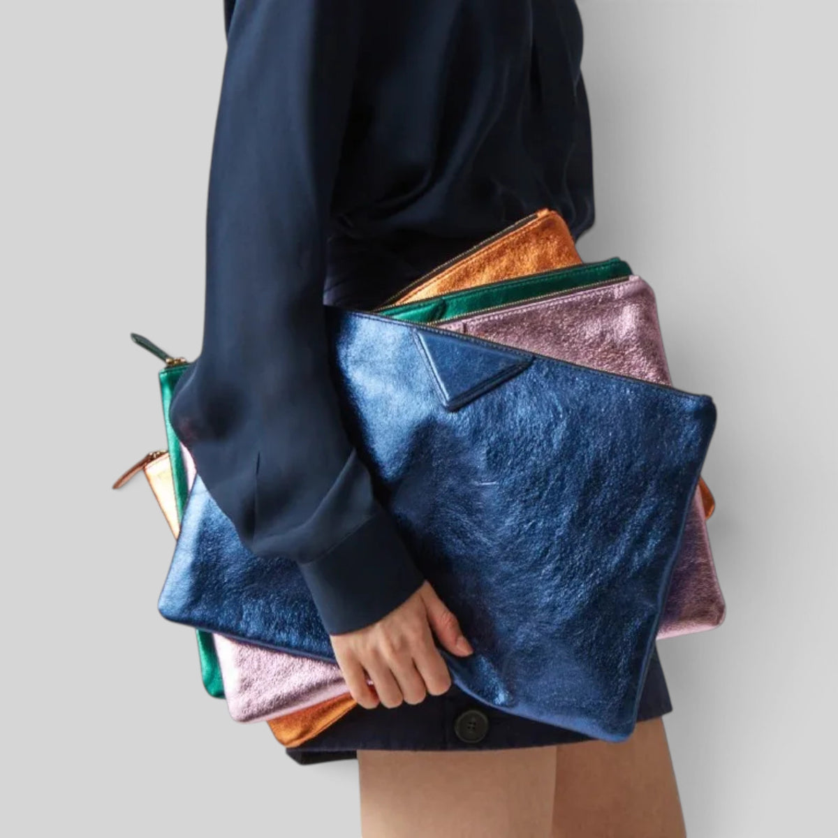 A person in a dark navy top holds a stack of metallic zip pouches in blue, pink, green, and orange against a light gray background.