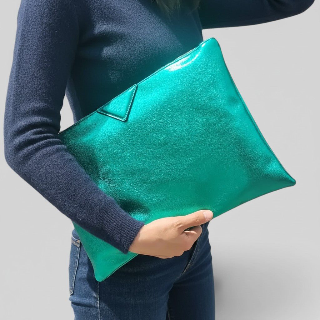 A person wearing a navy sweater and blue jeans holds a shiny teal-green envelope clutch bag against a plain light gray background.