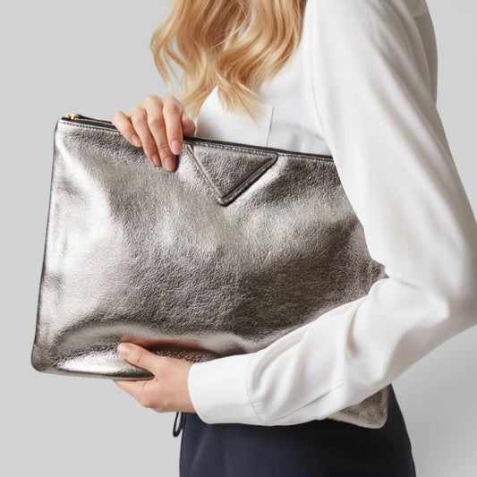 A woman in a white blouse holds a metallic silver clutch bag with a triangular front detail against a light gray background.