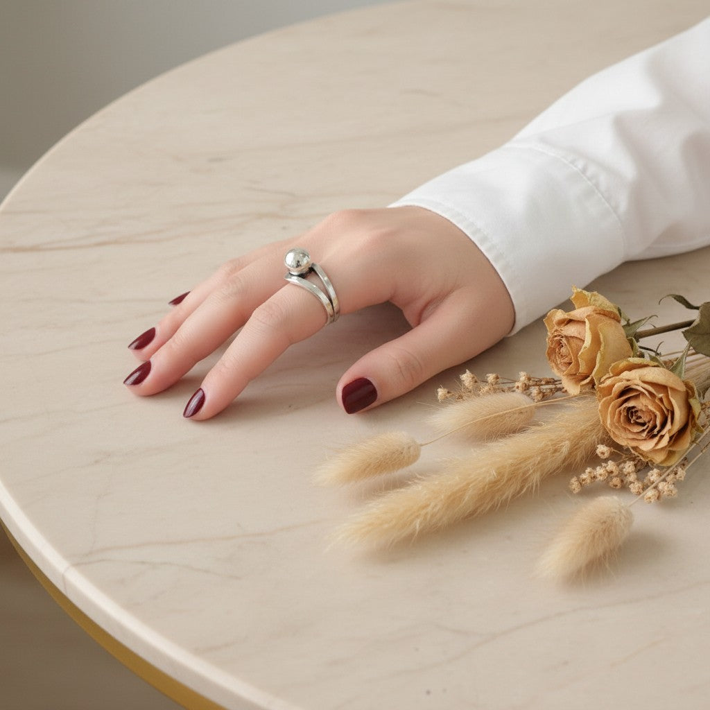 A silver ring with a small white flower and green leaves, worn on a woman's hand with dark burgundy nail polish, resting on a light beige marble table next to dried roses.