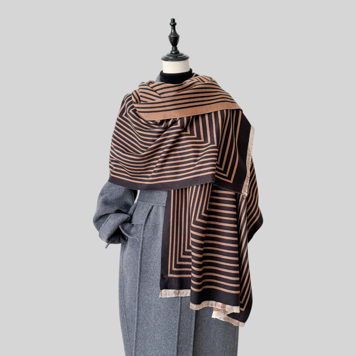 A patterned shawl draped over a gray coat on a mannequin torso, featuring geometric stripes in brown, beige, and black on a light gray background.