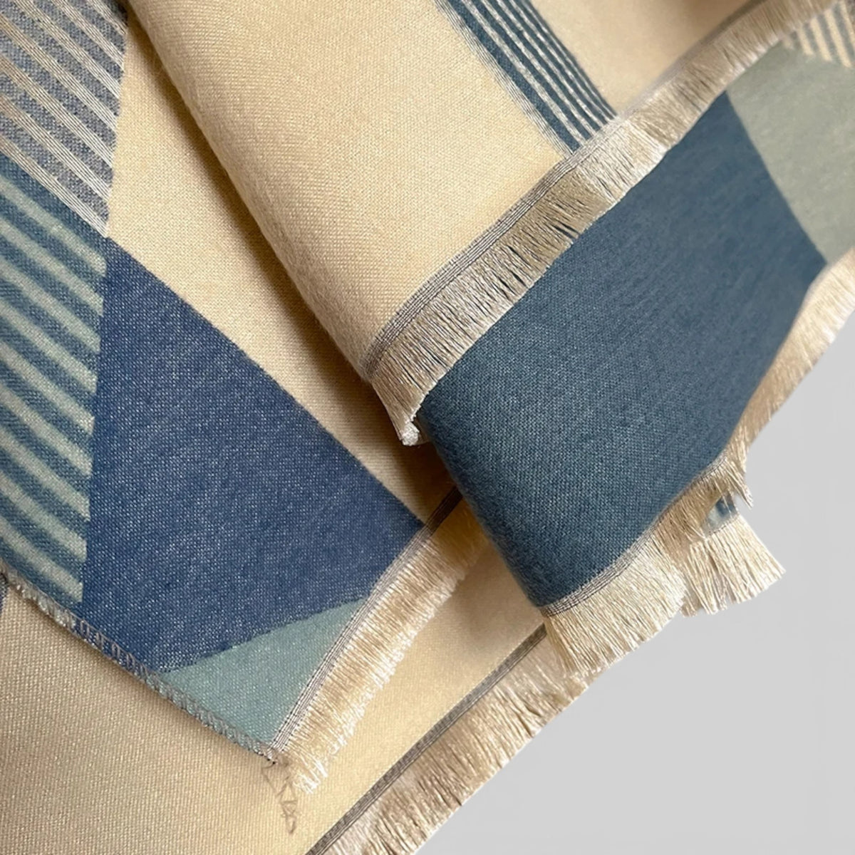A close-up of a woven scarf with a geometric pattern in cream, slate blue, and light sage green, finished with fringed edges.