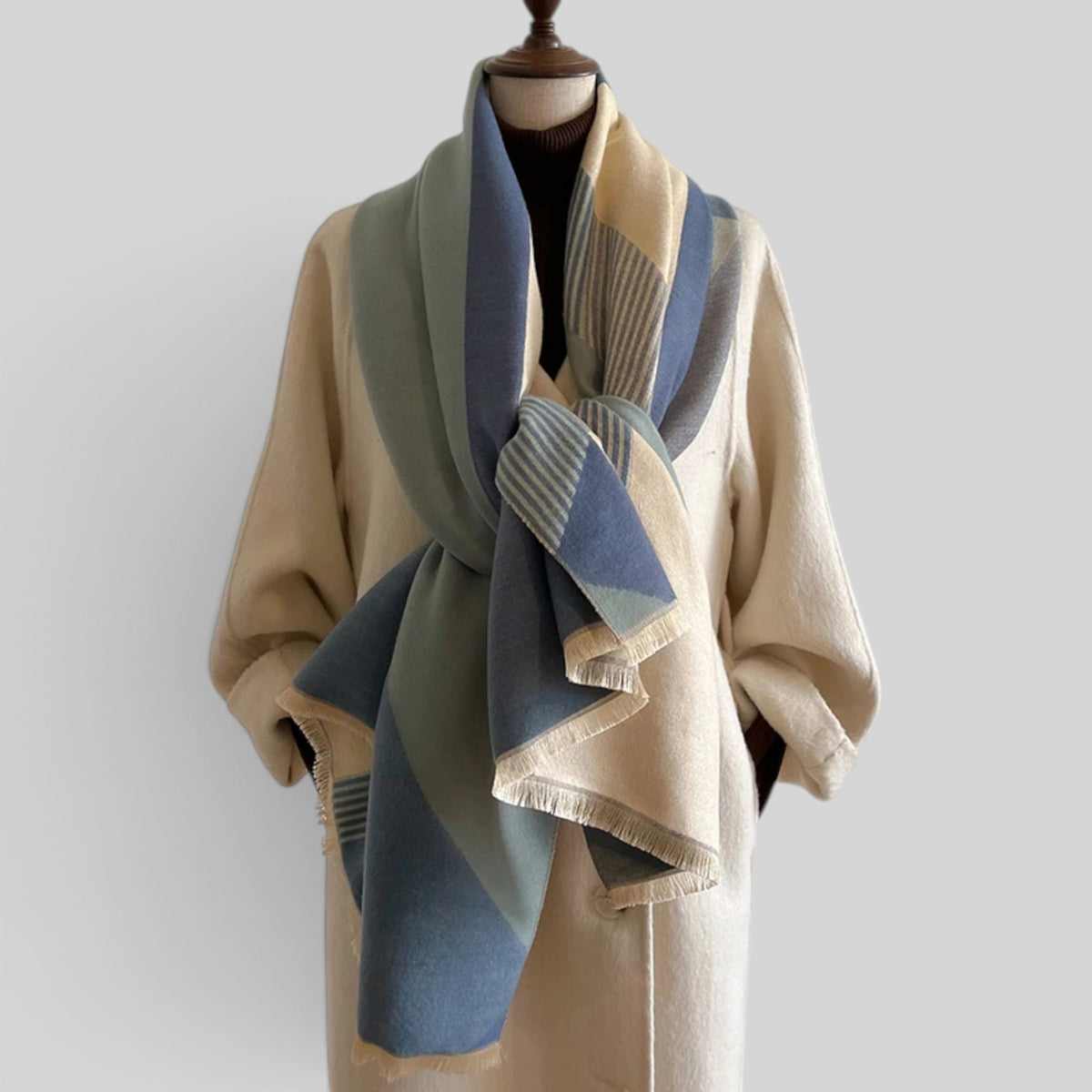A cream-colored wool coat displayed on a mannequin torso, styled with a large, fringed scarf in shades of blue, sage green, and cream.