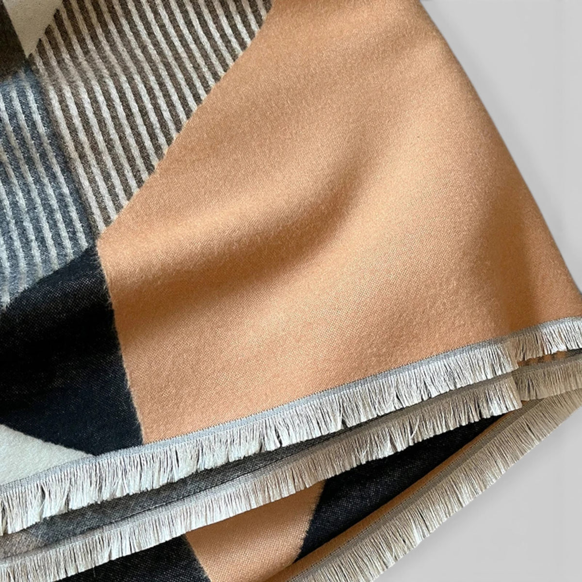 A close-up of a folded woven textile with a geometric color-block pattern in warm beige, charcoal gray, and off-white, finished with fringe trim.
