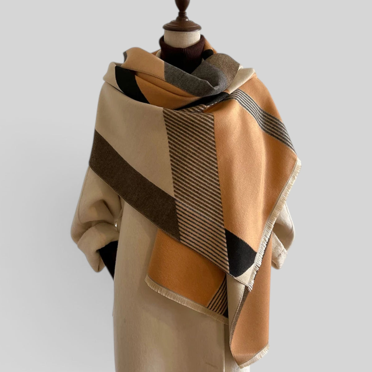 A large plaid shawl with a geometric pattern in beige, mustard yellow, brown, black, and gray, draped on a cream-colored mannequin torso against a solid light gray background.