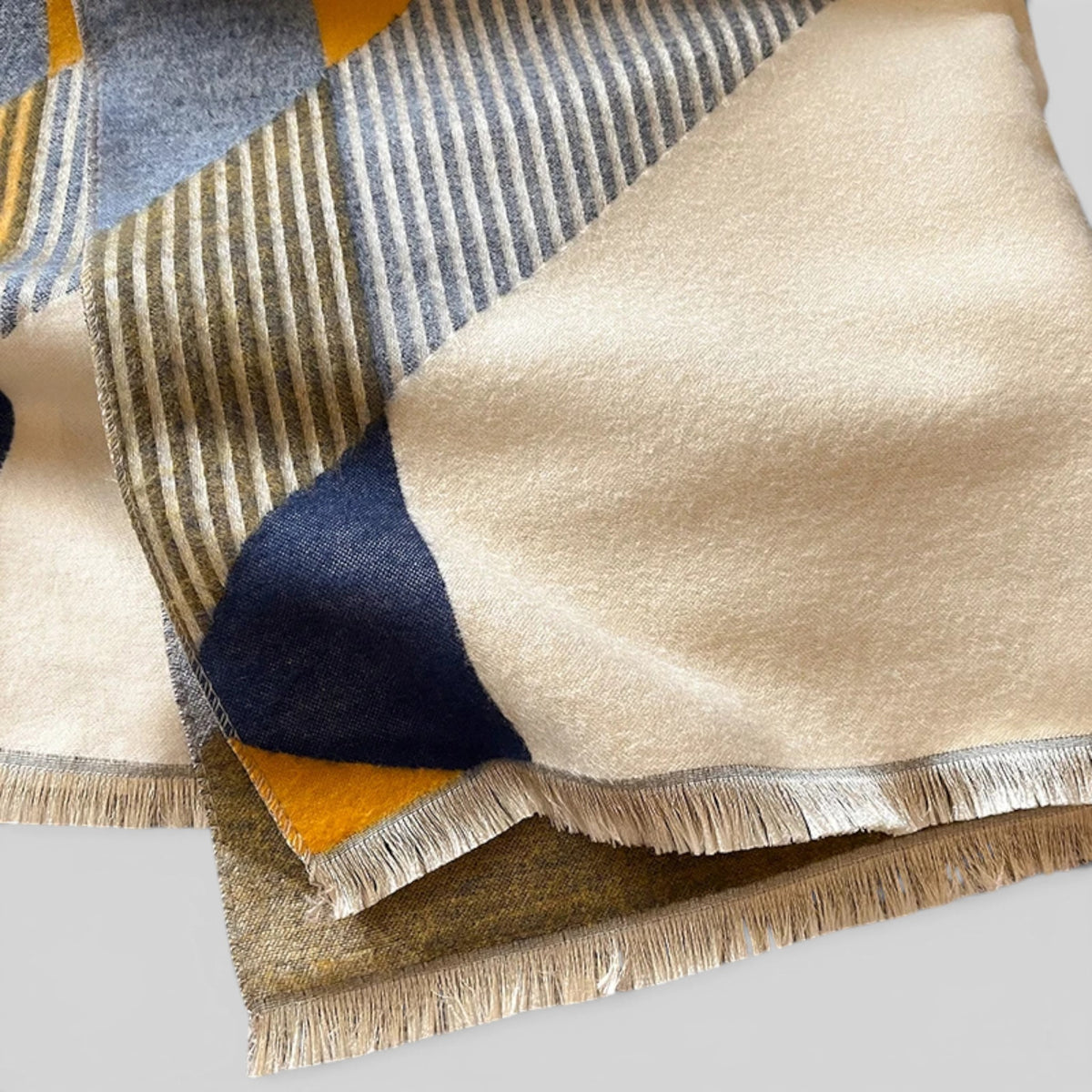 A close-up of a folded woven textile with a geometric patchwork pattern in cream, navy, mustard yellow, gray, and olive tones, finished with fringe along the edges.
