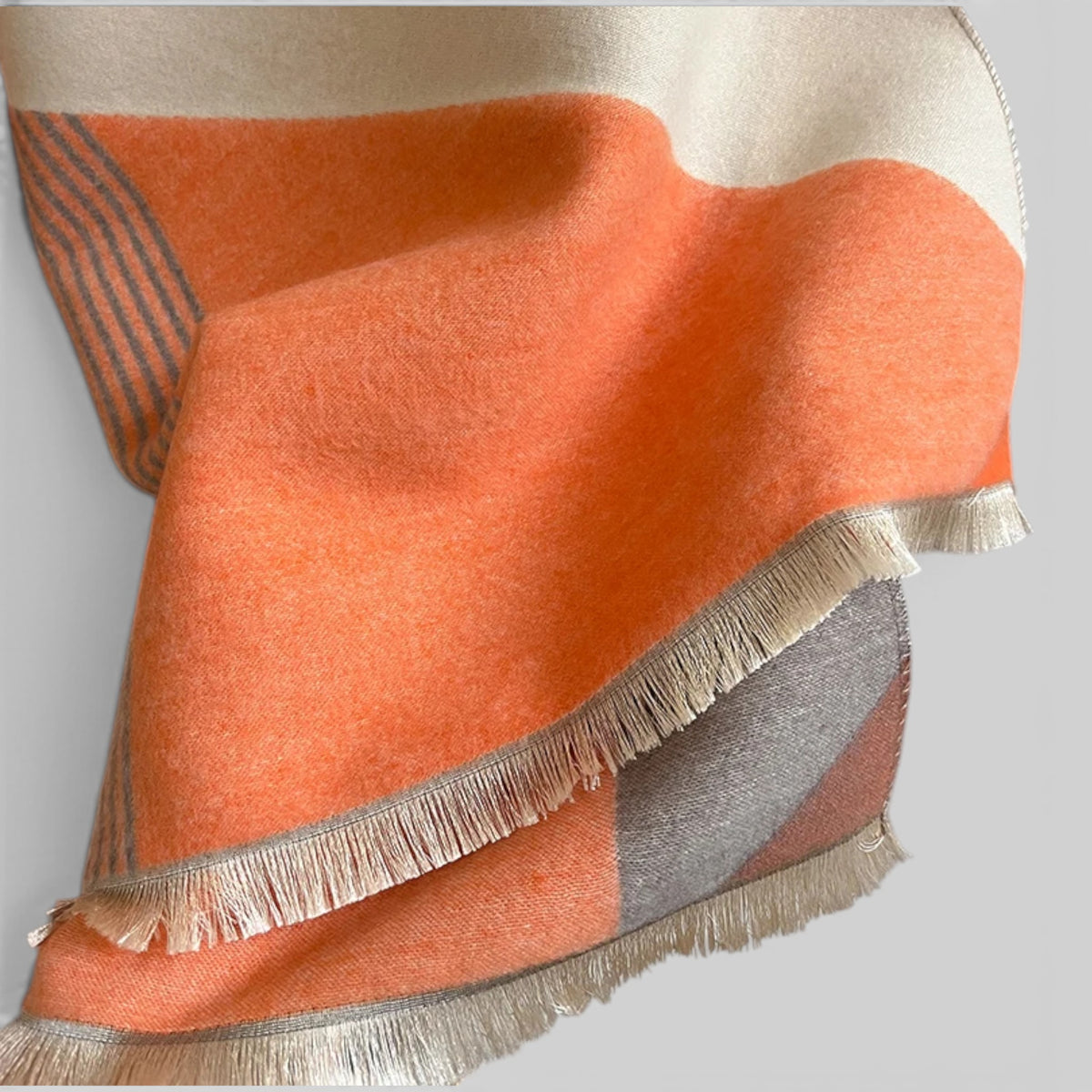 A close-up of a soft woven scarf with a color-blocked pattern in orange, cream, and gray, featuring cream fringe along the edges.