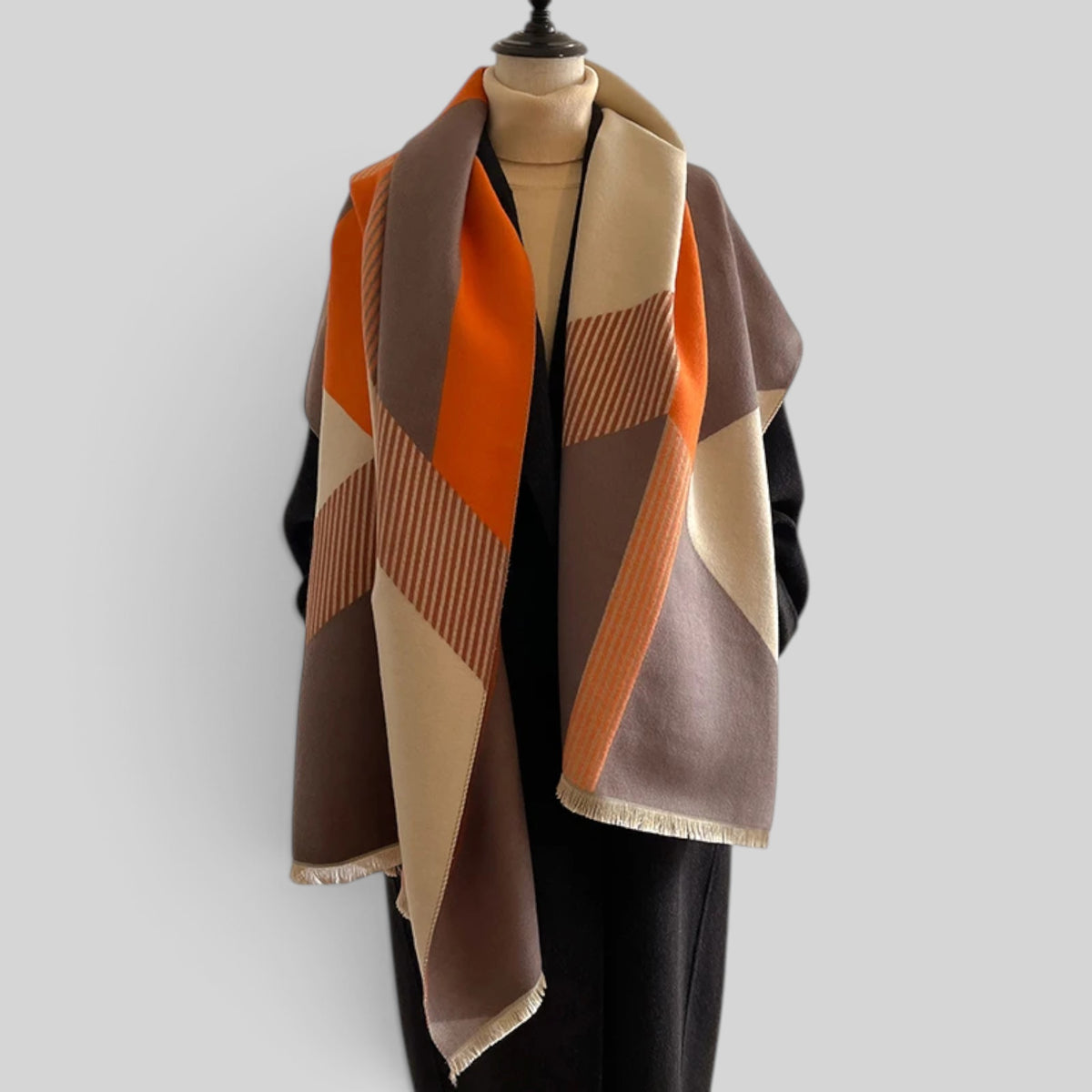 A large geometric patterned scarf draped over a black mannequin torso, featuring blocks of orange, beige, brown, and taupe with fringed edges, set against a solid light gray background.