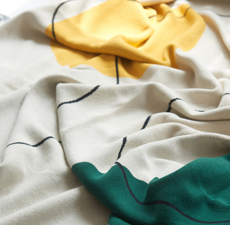 A close-up of a soft textile with a cream base, a large yellow abstract shape near the top, a teal green shape at the bottom, and thin black curved line accents.