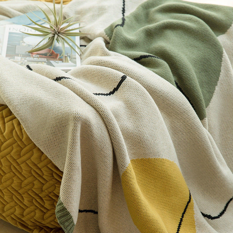 A close-up of a knitted throw blanket draped over a textured yellow pillow, featuring a cream base with a large mustard yellow geometric shape, a sage green section, and thin black stitched line accents. A small plant and magazine are visible in the background.