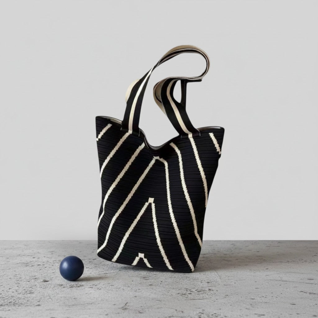 A black woven tote bag with diagonal cream stripes and a single shoulder strap, standing on a gray concrete surface next to a small dark blue sphere against a light gray background.