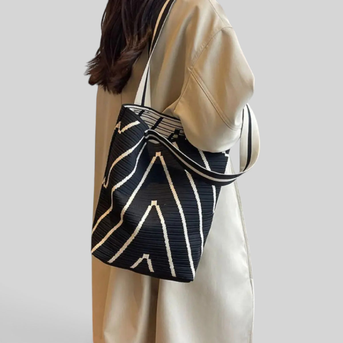 A person wearing a beige satin dress carries a black bucket-style shoulder bag with a cream geometric stripe pattern, shown against a solid light gray background.