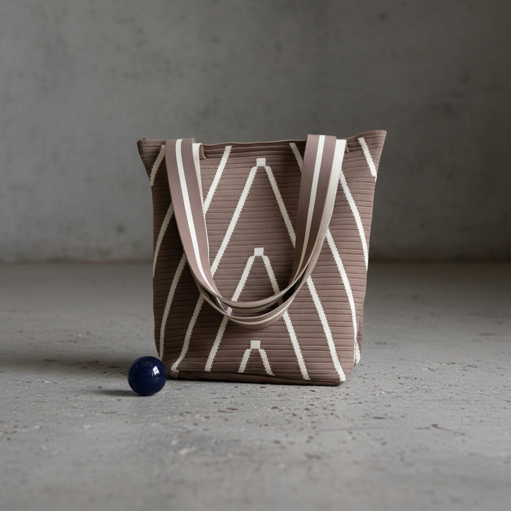A pleated tote bag in taupe brown with a cream geometric pattern and matching shoulder straps, standing on a concrete floor next to a small dark blue sphere.