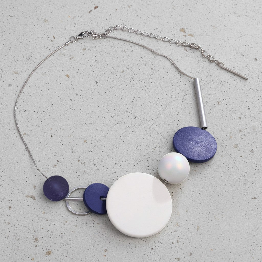 A modern statement necklace featuring a silver chain, a large white circular disc, a smaller white pearl bead, a textured metallic blue disc, a flat blue disc, and a small dark purple disc, displayed on a speckled light gray surface.