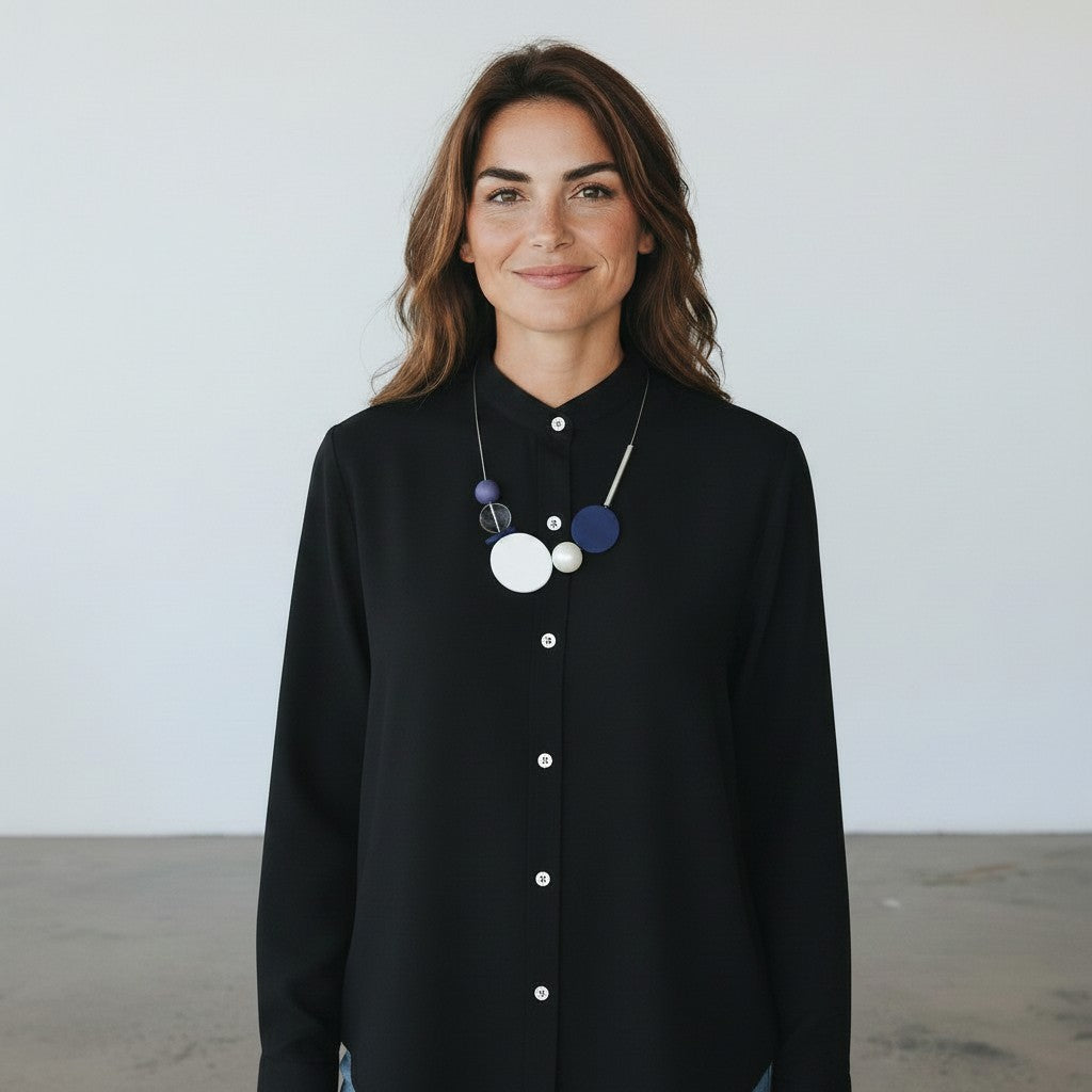 A woman with brown hair wearing a black long-sleeve button-up shirt and a chunky multi-colored geometric pendant necklace, standing against a plain light gray wall.