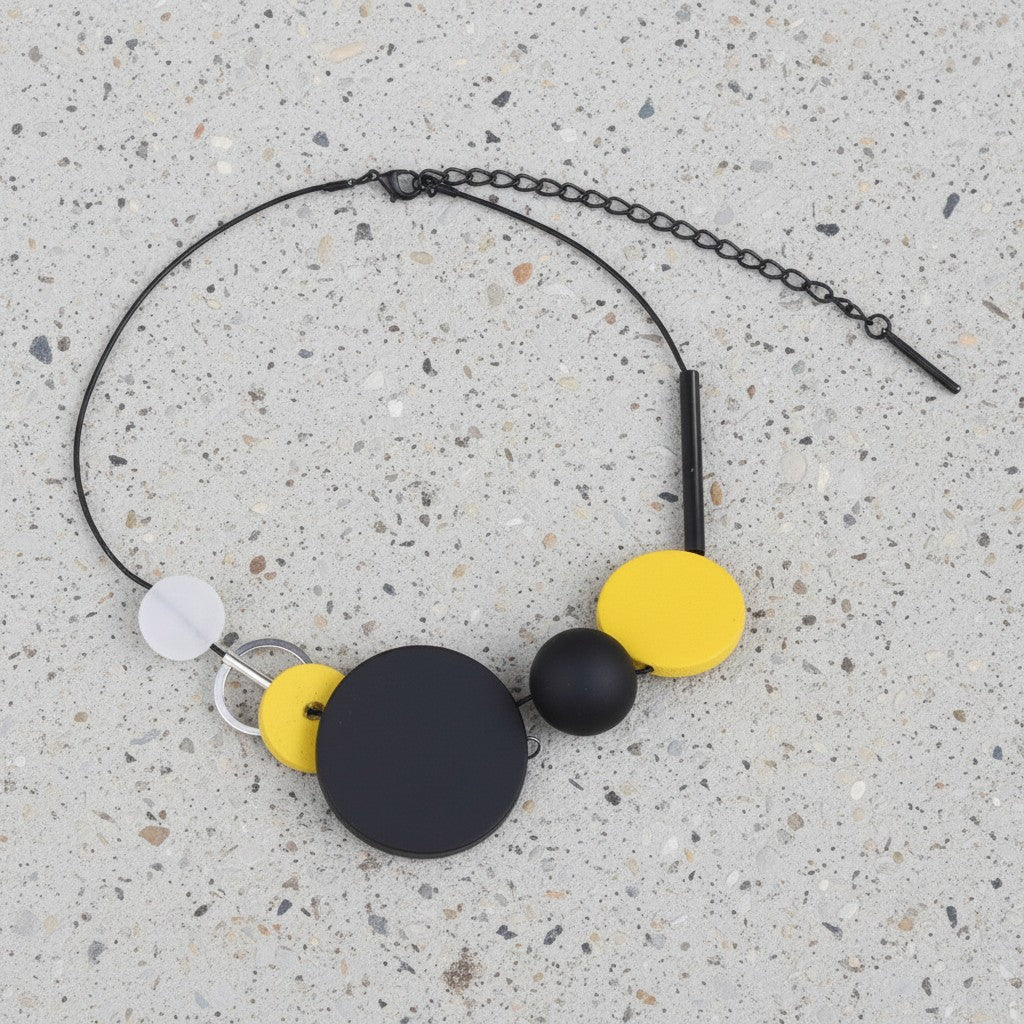 A modern beaded necklace with a black chain, featuring matte black and bright yellow circular beads, a small black sphere, and a white disc, laid flat on a speckled gray concrete surface.