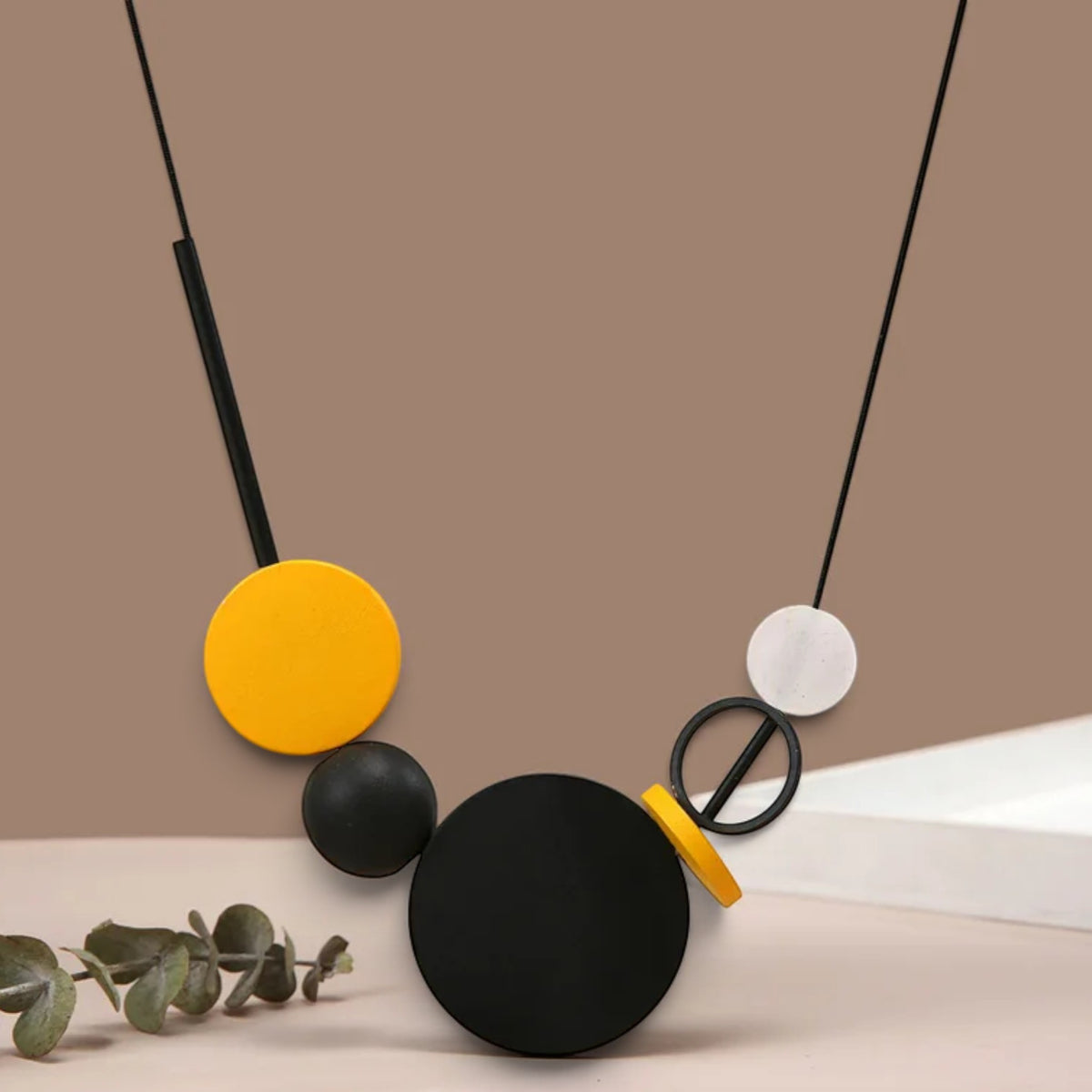 Abstract geometric pendant necklace with matte black, yellow, and white circular and oval elements on black cords, styled on a beige surface with a brown background and a sprig of eucalyptus.