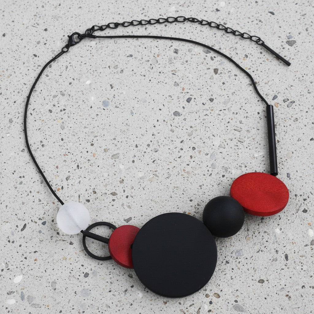 A modern abstract statement necklace featuring matte black and red circular discs, a translucent white disc, and a black oval bead on a black chain, displayed on a speckled concrete surface.