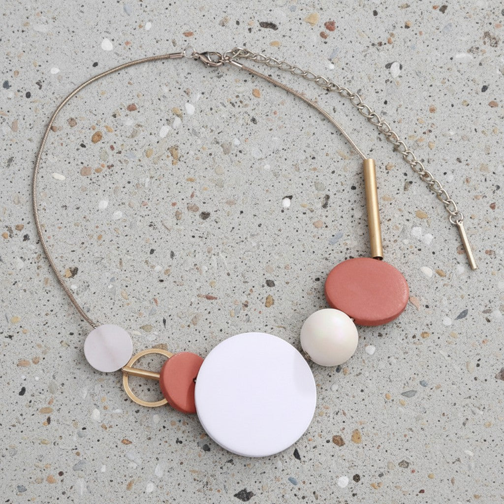 A bohemian-style necklace featuring a silver-tone chain, a large white circular disc, terracotta and cream discs, and gold-tone metal accents, displayed on a speckled concrete surface.