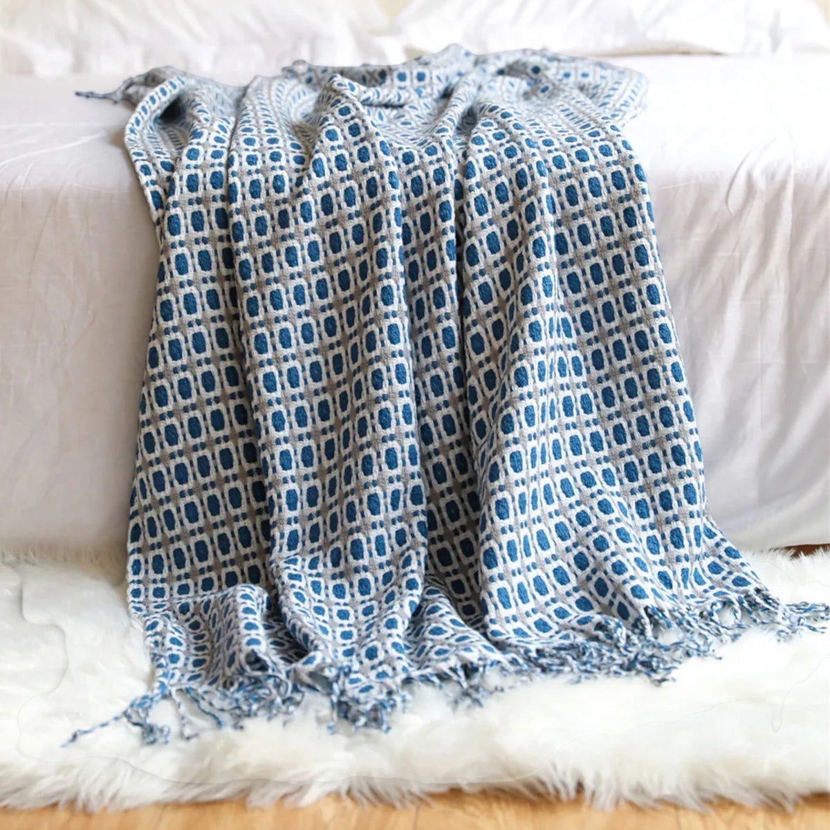 A blue and white patterned throw blanket with fringed edges draped over a white bed on a fluffy white rug.