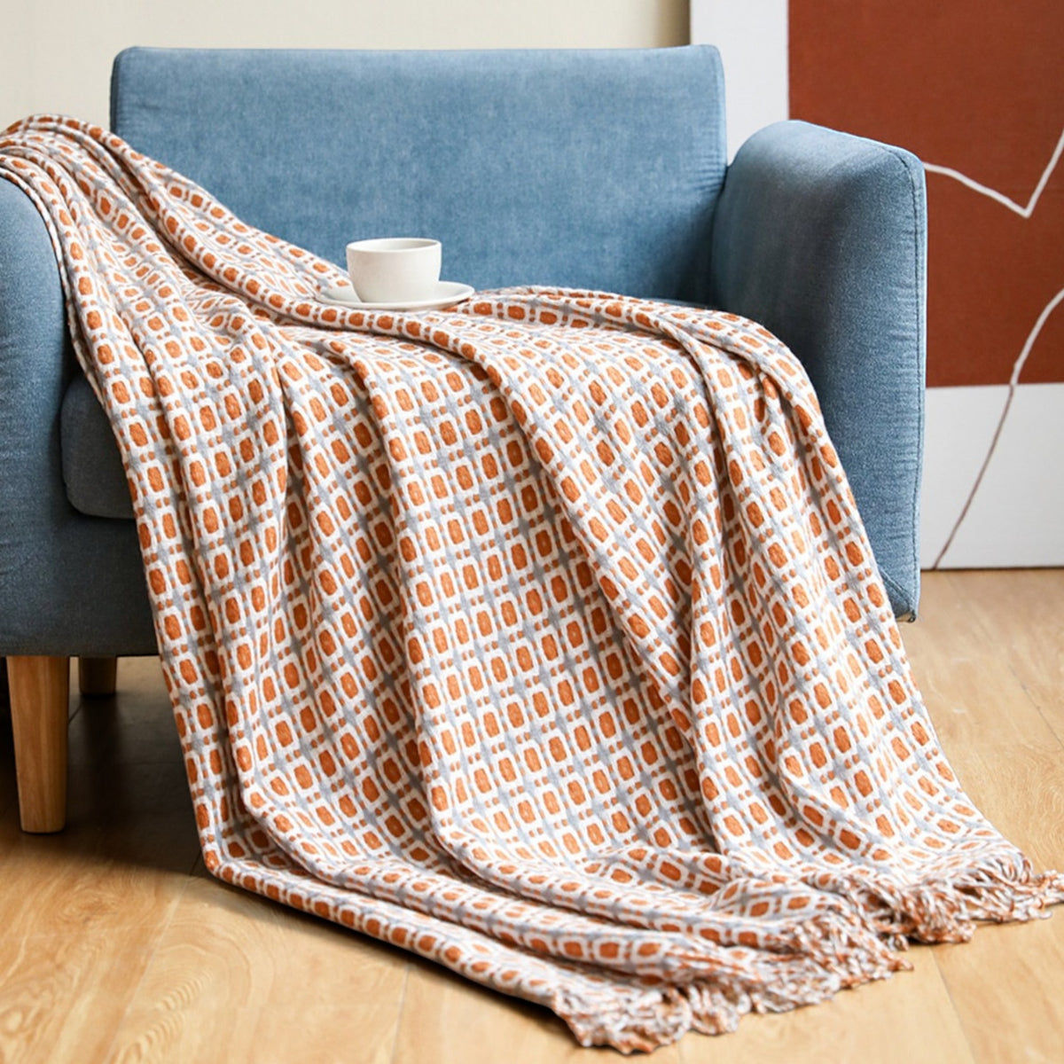 A patterned throw blanket draped over a blue armchair in a living room setting, with a white cup on top. The blanket features a geometric orange and gray pattern on a cream background.