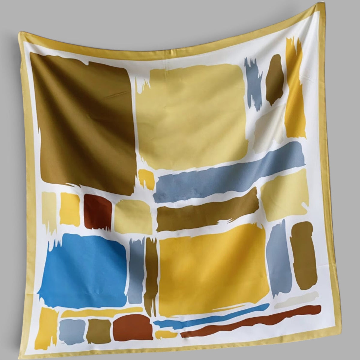 A square silk scarf with an abstract geometric pattern of paint blocks in yellow, blue, brown, and gray, finished with a gold border, shown against a neutral gray background.