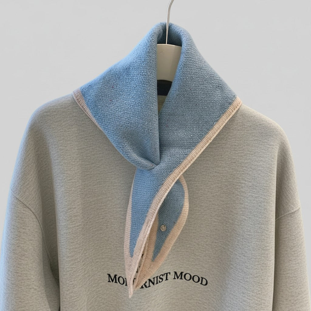A light gray oversized knit sweater with a wide collar, featuring a light blue knit neck scarf with beige trim and black text reading 'MODERNIST MOOD' on the chest, displayed on a hanger against a light gray background.