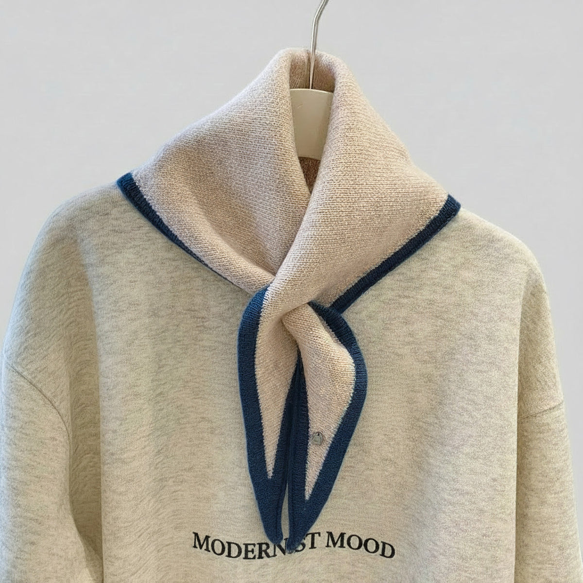 A cream-colored knit sweater with a wide folded collar, featuring dark blue trim along the collar edge and the words 'MODERNIST MOOD' printed in black across the chest. The sweater is displayed on a hanger against a light gray background.
