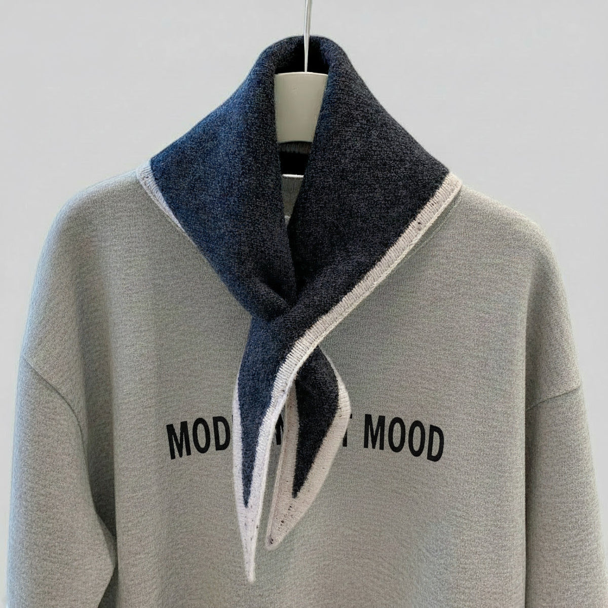 A light gray knit sweater with black 'MOOD' text on the chest, styled with a dark navy blue and white striped scarf on a hanger against a light gray background.