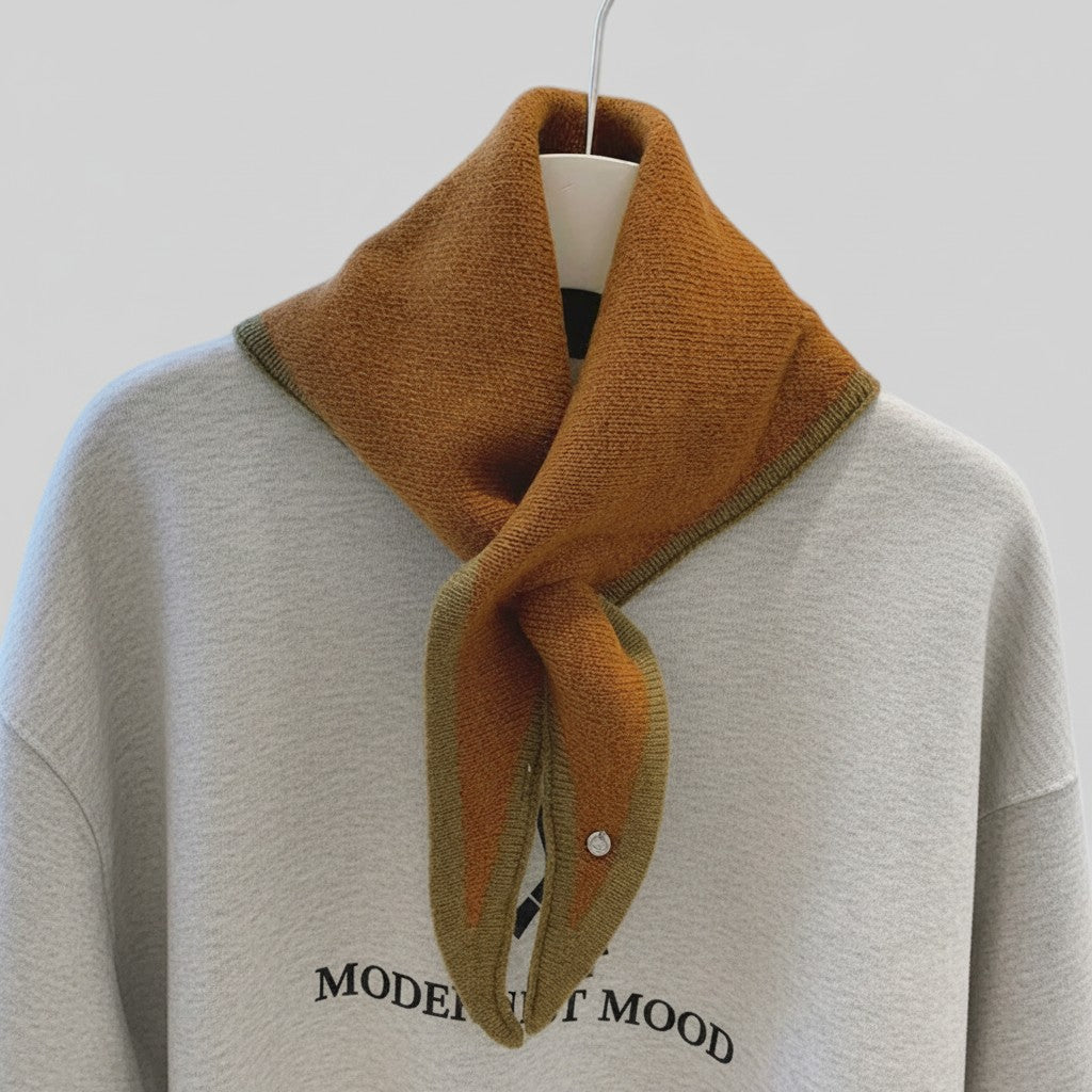 A mustard yellow and olive green knit scarf draped around the collar of a light gray sweatshirt on a hanger, against a plain light gray background.