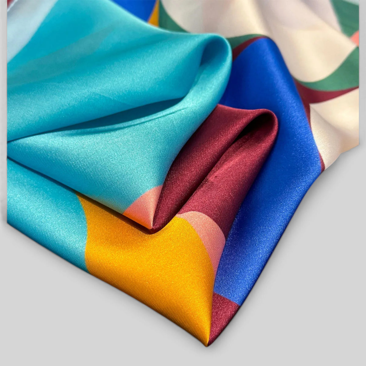 A folded silk scarf with a vibrant geometric color-block pattern, featuring shades of turquoise, royal blue, mustard yellow, burgundy, and cream on a light gray background.